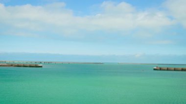Güneşli bir günde Dunkirk limanından çıkarken açık deniz manzarası. Turkuaz su mavi gökyüzünün altında liman duvarları arasında uzanıyor. Sakin okyanus ufku Fransız sahil limanının kapısında beliriyor.