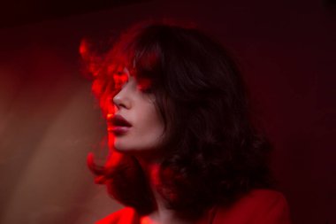 Makyajlı ve saç stili olan genç bir kadının yakın plan portresi, kırmızı takım elbise, kapalı gözler, kırmızı stüdyo ışığı ve efektleri..