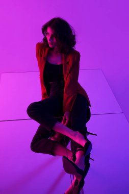 Kırmızı takım elbiseli, esmer, şehvetli bir mankenin tam boy fotoğrafı. Neon ışıklı siyah kotlar, yerde aynada poz veriyor..