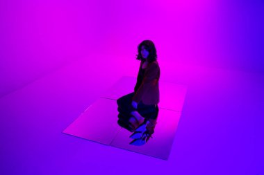 Kırmızı takım elbiseli çekici esmer bir mankenin tam boy fotoğrafı. Siyah kotlu, karışık pembe ve mor neon ışıklı, yerde aynaya poz veren..