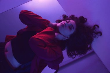 Kırmızı takım elbiseli esmer, siyah kotlu, pembe ve mor ışıklı genç bir mankenin aynaya uzanmış görüntüsü. Kavramsal moda fotoğrafı.