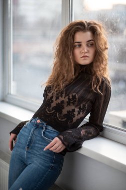 Profile portrait of a gorgeous young woman with curly hair, wear black blouse and jeans, posing near bright windows.