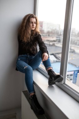 Full length image of a gorgeous young woman with curly hair, wear black blouse and jeans, posing near windows, looking at the side.