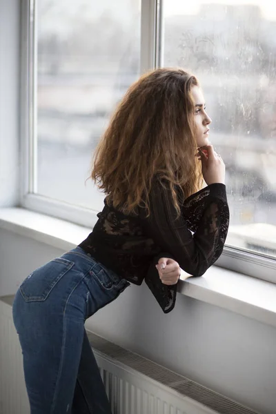 Gorgeous young woman with curly hair, wear black blouse and jeans, posing and looking at the window.