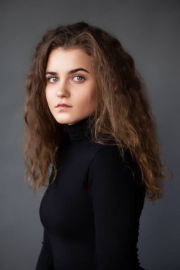 Front image portrait of a girl with curly hair and natural makeup, dressed in a black , on a grey background.