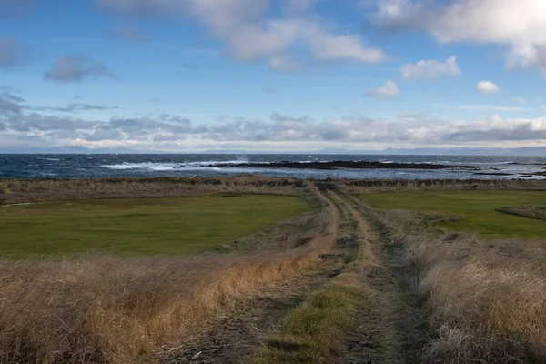 Country road in between  fields and pastures. Horizon of the Icelandic sea (Atlantic ocean) in the background. Blue sky with white clouds in the autumn. Holmsvollur, Iceland.