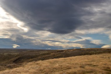 Late afternoon country with a dark brown soil and dry grass and plants in the autumn. Horizon of the mountains in the background. Intense clouds on the sky. Reykhold, south-middle part of Iceland.
