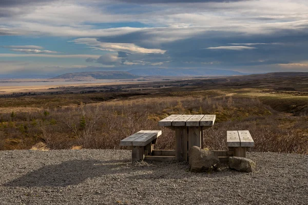 Country with trees, bushes, grass. Mountains on the horizon. Natural wooden desk and benches to sit, relax, eat and enjoy the panorama. Intense clouds on the sky. Reykhold, south-middle Iceland