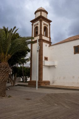 White facade of a church, with orange-brown details. Tower with a belfry. Church located in a park with trees, bushes and plants, with a lot of place to relax. Antigua, Fuerteventura, Canary Islands, Spain.