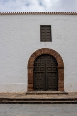 White facade of the church.Side gate with a stone frame. Decorative structure of a wooden closed gate and a window above. Line of the roof and cloudy sky. Antigua, Fuerteventura, Canary Islands, Spain