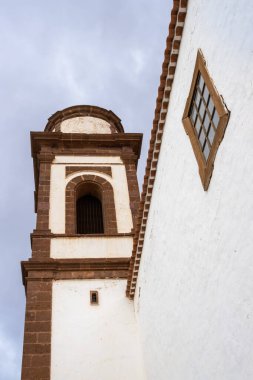 Tower of a church with belfry. White facade and brown details. Window with a wooden brown frame. Line of the roof. Cloudy sky. Antigua, Fuerteventura, Canary Islands, Spain.