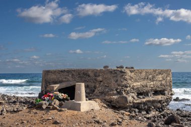 Bunker on the east coast of Atlantic ocean (Africa side). Still vivid memory, many artificial flowers. Atlantic ocean and blue sky with white clouds. Fuerteventura, Canary Islands, Spain.
