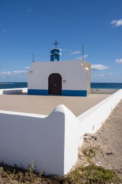 Small church on the east coast of the Atlantic ocean named Ermita de la Virgen del Pino. Simple combination of white and blue. Belfry and a cross. Puerto Lajas, Fuerteventura, Canary Islands.