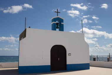 Small church on the east coast of the Atlantic ocean named Ermita de la Virgen del Pino. Simple combination of white and blue. Belfry and a cross. Puerto Lajas, Fuerteventura, Canary Islands.