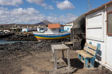 Koyda renkli bir balıkçı teknesi. Güneşli kış günü. Sahilde bankı ve masası olan ahşap bir ev. Beyaz bulutlu mavi gökyüzü. Arka plandaki dağ. Playa del Jablito, Fuerteventura