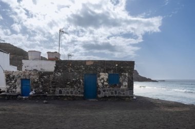 Doğrudan Atlantik Okyanusu 'nun kıyılarına inşa edilmiş, koyu mavi renkli taşlardan yapılmış bir hosue. Arka plandaki dağ. Büyük beyaz bulutlu mavi gökyüzü. Los Molinos, Fuerteventura.