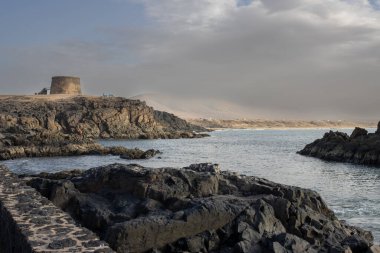 Atlantik Okyanusu körfezi, kayalar ve kayalıklarla kaplı. Tepedeki tarihi taş kule. Kış mevsiminde bulutlu bir gökyüzü. El Cotillo, Fuerteventura, Kanarya Adaları, İspanya.