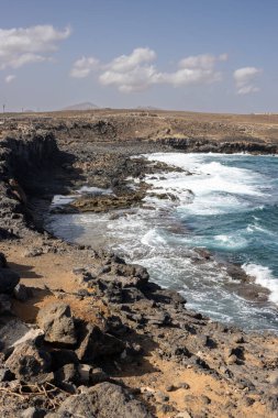 Atlantik Okyanusu kıyısındaki koyu kahverengi kayalar. Kış mevsiminde açık bulutlu mavi gökyüzü. Güneşli bir gün. Piscina Doğal, Castillo Caleta de Fuste, Fuerteventura, Kanarya Adaları.