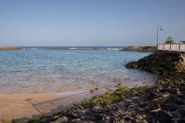 Atlantik okyanusunun sakin suları. Sağ taraftaki taşlar, üzerinde mavi bir sokak lambası olan bir köprünün tabanı.