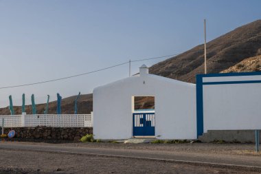 Dağın altındaki bir binaya giriş. Giriş ve çitin beyaz cephesi. Parlak mavi detaylar. Mavi gökyüzü. Pozo Negro, Fuerteventura, Kanarya Adaları, İspanya.