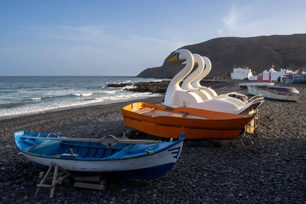 Stock image Pebble beach in the winter season. Fishing wooden boats and swans water bikes. Houses and Mountain in the background. Small waves of Atlantic ocean and blue sky. Pozo Negro, Fuerteventura