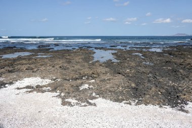 Atlantik Okyanusu sahilindeki karanlık kayalar. Patlamış mısır şeklinde kırık mercan parçalarıyla dolu beyaz alanlar. Açık beyaz bulutlu mavi gökyüzü. Playa del Bajo de la Burra ve Playa el Mejilon, Fuerteventura.