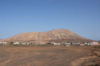 Taşlı ve taşlı kum toprağı. Arka plandaki dağ. Tepenin altındaki köyün evleri. Parlak mavi gökyüzü. Villaverde, Fuerteventura, Kanarya Adaları, İspanya.