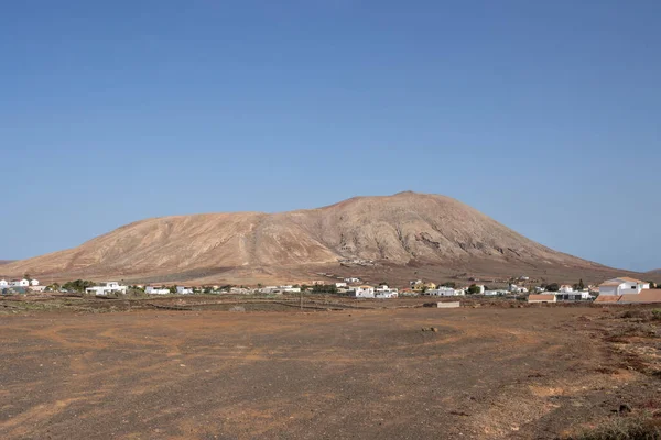 stock image Sandy soil with stones and rocks. Mountain in the background. Houses of the village under the hill. Bright blue sky. Villaverde, Fuerteventura, Canary Islands, Spain.