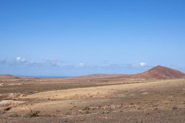 Adanın merkezindeki bir yaylanın etrafındaki kuru volkanik kayalık dağlar. Arka planda Atlantik Okyanusu. Beyaz bulutlu mavi gökyüzü. Merkez Fuerteventura, Kanarya Adaları, İspanya.