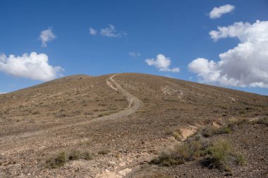 Adanın merkezindeki bir yaylanın etrafındaki kuru volkanik kayalık dağlar. Taşra yolu. Beyaz bulutlu mavi gökyüzü. Merkez Fuerteventura, Kanarya Adaları, İspanya.