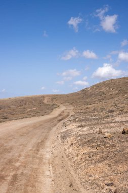 Adanın merkezindeki bir yaylanın etrafındaki kuru volkanik kayalık dağlar. Taşra yolu. Beyaz bulutlu mavi gökyüzü. Merkez Fuerteventura, Kanarya Adaları, İspanya.