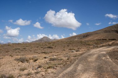 Adanın merkezindeki bir yaylanın etrafındaki kuru volkanik kayalık dağlar. Taşra yolu. Beyaz bulutlu mavi gökyüzü. Merkez Fuerteventura, Kanarya Adaları, İspanya.