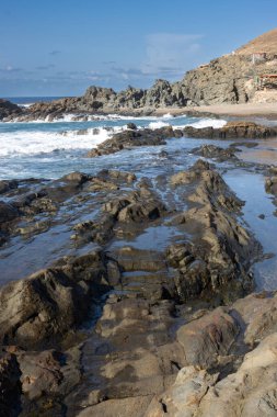 Adanın batısındaki Atlantik Okyanusu plajı. Koyu kahverengiden siyah kayalara doğal havuzlar oluşturur. Kışın güneşli bir gün. Aguas Verdes, Fuerteventura, Kanarya Adaları, İspanya.