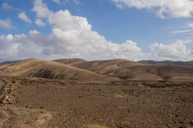Kuru volkanik arazi ve arka plandaki dağlar, adanın batısında. Beyaz bulutlu mavi gökyüzü. Yürüyüş için yer var. Gambuesa de Llano del Sombrero, Betancuria, Fuerteventura, Kanarya Adaları.