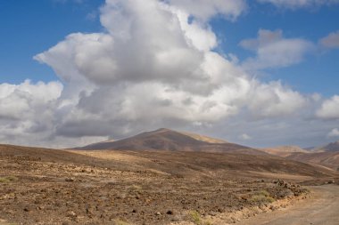 Adanın batısındaki kuru volkanik dağlar. Taşra yolu. Beyaz bulutlu mavi gökyüzü. Gambuesa de Llano del Sombrero, Betancuria, Fuerteventura, Kanarya Adaları, İspanya.
