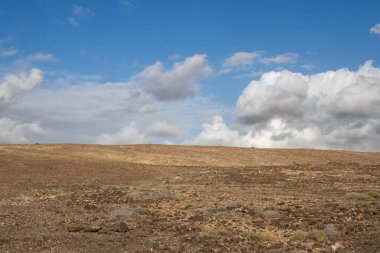 Kışın, adanın batısında birçok taş, kaya ve bazı bitkilerin bulunduğu kuru topraklar. Beyaz bulutlu parlak mavi gökyüzü. Gambuesa de Llano del Sombrero, Betancuria, Fuerteventura, İspanya