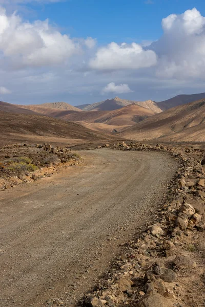 Dry volcanic mountains in the west of the island. Country gravel road. Blue sky with white clouds. Gambuesa de Llano del Sombrero, Betancuria, Fuerteventura, Canary Islands, Spain.