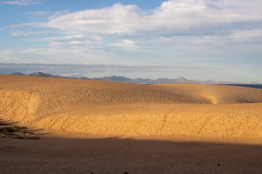 Doğal parktaki kum tepeleri, altın saat boyunca renkliydi. Arka planda Lanzarote Dağları var. Genişliğinde bulutlar olan mavi gökyüzü. Park Natural de Dunas de Corralejo, Fuerteventura, İspanya.