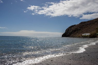 Atlantik Okyanusu 'nun sakin suları ve küçük şehirde kış mevsiminde sakin bir ortam. Sahilin kıvrımları bir dağla bitti. Beyaz bulutlu mavi gökyüzü. Giniginamar, Fuerteventura, İspanya.