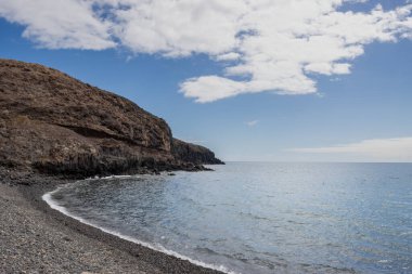 Atlantik Okyanusu 'nun sakin suları ve küçük şehirde kış mevsiminde sakin bir ortam. Sahilin kıvrımları bir dağla bitti. Beyaz bulutlu mavi gökyüzü. Giniginamar, Fuerteventura, İspanya.