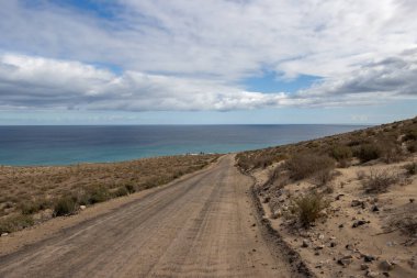 Tepeden aşağı çakıl yolu, Atlantik Okyanusu yönünde. Beyaz bulutlu mavi gökyüzü. Costa Calma, Fuerteventura, Kanarya Adaları, İspanya.