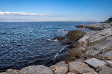 Denizin küçük dalgalı mavi rengi. Beyaz bulutlu mavi gökyüzü. Ön planda kıyı kayaları var. Limenas Thasou, Thassos (Tassos) adasının başkenti, Yunanistan.