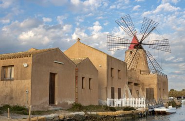 Deniz tuzu işlemek için tuzluk ve yel değirmenleri. Aralarında kanal var. Baharda beyaz bulutlu mavi gökyüzü. Lagoon dello Stagnone, Marsala, Sicilya, İtalya.