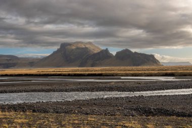 Ot ve yosun rengi sarı-yeşil bir ülke. Bir nehir çizgisi ve arka planda bir dağ. Sonbaharda bulutlu ve dramatik bir gökyüzü. Onundarhorn, Güney İzlanda.