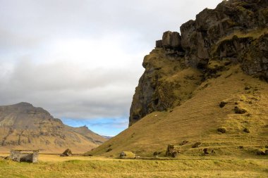 Çimlerin ve yosunların sarı-yeşil rengi, tüm zemini ve dağı kaplıyor. Sonbaharda bulutlu bir gökyüzü. Atlantik Okyanusu 'nun güneyinde, Kvernufoss, Güney İzlanda yakınlarında..