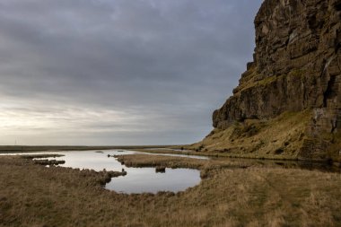 Yüksek dağ çimlerle ve yosunlarla kaplı ve yanında bir göl var. Sudaki yansıma. Bulutlu sonbahar havası. Kvernufoss bölgesi, güney-merkez İzlanda.