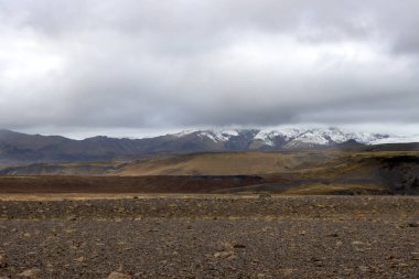 Öndeki volkanik çakılların koyu kahverengi rengi. Arka planda dağlar var, tepesinde kar var. Sonbaharda yoğun bulutlu bir gökyüzü. Güney-Orta İzlanda.