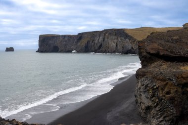 Atlantik okyanusunu çevreleyen kayalar. Bulutlu gökyüzü, bulutların arasında güneş ışığı var. Okyanusun sakin sularında siyah kayalar. Dyrholaey, Güney İzlanda.