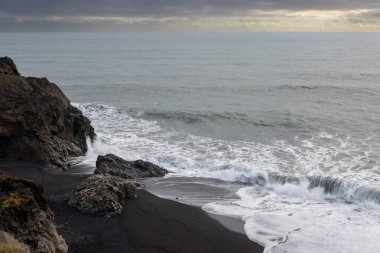 Atlantik okyanusunu çevreleyen kayalar. Bulutlu gökyüzü, bulutların arasında güneş ışığı var. Okyanusun sakin sularında siyah kayalar. Dyrholaey, Güney İzlanda.