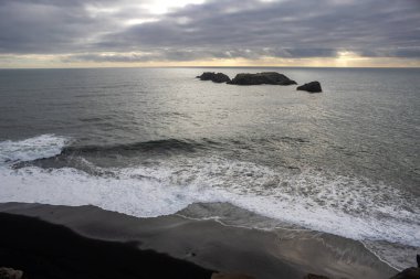 Atlantik Okyanusu ve bulutlu bir gökyüzü, bulutların arasında güneş ışığı. Okyanusun sakin sularında siyah kayalar. Dyrholaey, Güney İzlanda.